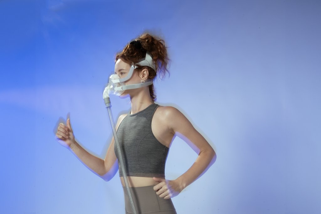 A curly haired woman wearing yoga attire, running in place in front of a blue background with a VO2 Max Training Mask on her face. 