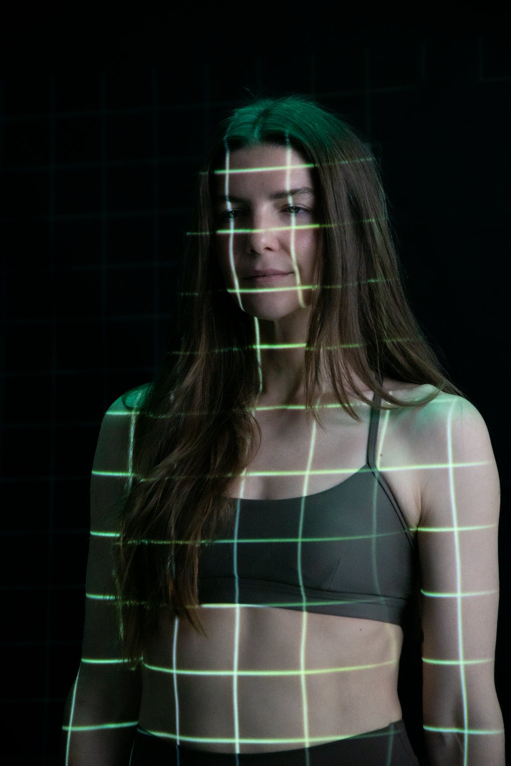 A woman in a sports bra and tank top stands confidently in front of a green screen background. She is facing the camera, her gaze intense. Her body is covered in the a gird of green light from a full body scan. 